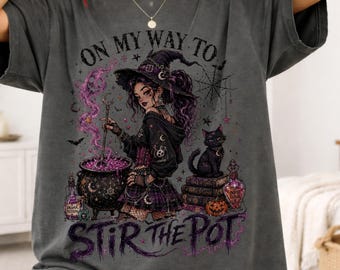 May include: A charcoal gray t-shirt featuring a graphic print of a witch stirring a cauldron with a black cat, books, and potion bottles. The text reads ON MY WAY TO STIR THE POT. The design includes purple smoke, stars, and a spiderweb.