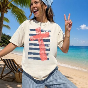 May include: A cream-colored short-sleeve t-shirt featuring a graphic print of a distressed, coral-colored cross layered over horizontal navy blue brushstroke stripes. The shirt has a crew neckline and a relaxed fit, made of a solid-colored fabric.
