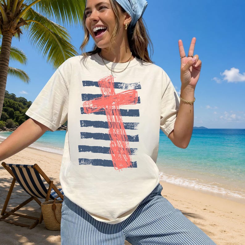 May include: A cream-colored short-sleeve t-shirt featuring a graphic print of a distressed, coral-colored cross layered over horizontal navy blue brushstroke stripes. The shirt has a crew neckline and a relaxed fit, made of a solid-colored fabric.