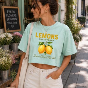 Op de afbeelding: Een licht mintgroen cropped T-shirt met een afbeelding van twee gele citroenen aan een tak met groene bladeren. De tekst LEMONS SUN RIPENED en Fresh Citrus Market staat in gele en groene letters in het midden op de voorzijde van het kledingstuk.