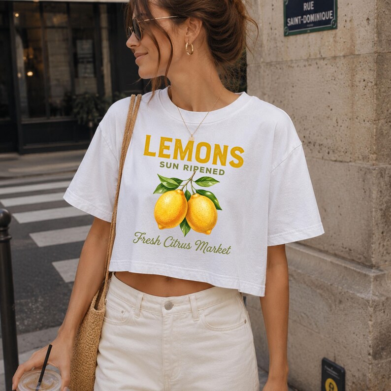 Op de afbeelding: Een wit kort T-shirt met een afbeelding van twee gele citroenen met groene bladeren. De tekst luidt LEMONS SUN RIPENED in het geel en Fresh Citrus Market in het groen. Het shirt heeft korte mouwen en een ronde hals, gedragen met een witte broek.