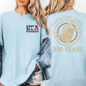 A woman wearing a light blue t-shirt with the text "USA freedom" and "Founding Fathers' Legacy" printed on it, along with a graphic of a feather quill and a document.