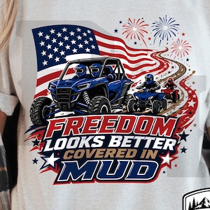 Puede incluir: Una camiseta gris claro con un gráfico de un UTV azul y dos ATV en un camino de tierra, con una bandera estadounidense y fuegos artificiales de fondo. El texto dice FREEDOM LOOKS BETTER COVERED IN MUD, con el logo RAISED ON MUD CO. abajo.