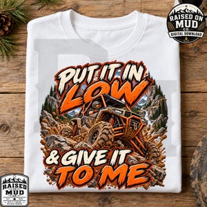 Könnte beinhalten: Ein weißes T-Shirt mit einer Grafik eines orangefarbenen Geländewagens, der durch felsiges Gelände fährt. Der Text "PUT IT IN LOW & GIVE IT TO ME" ist in fetten orangefarbenen Buchstaben gedruckt. Das Design zeigt Berge, Bäume und Schlammspritzer.