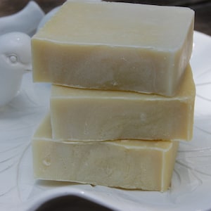 May include: Three rectangular bars of light-colored soap are stacked on a white, leaf-shaped dish. The soap has a slightly textured surface and a pale yellow hue. A white ceramic bird figurine is visible on the left side of the dish.