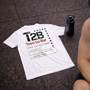 Könnte beinhalten: Weißes T-Shirt mit dem Text "T2B Toes-to-Bar" in Schwarz und Rot. Das Shirt enthält eine Definition der Übung und eine Reihe schwarzer Sterne. Eine schwarze Wasserflasche ist im Hintergrund zu sehen.