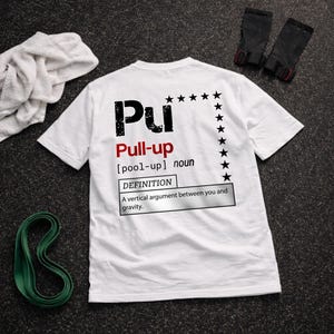 Könnte beinhalten: Weißes T-Shirt mit dem Wort "Pu" in einer verwaschenen schwarzen Schrift, gefolgt von "Pull-up" in Rot. Die Definition von Pull-up ist unten abgedruckt. Schwarze Sterne sind auf der rechten Seite des Shirts aufgedruckt. Ein weißes Handtuch, ein grünes Widerstandsband und schwarze Trainingshandschuhe sind ebenfalls sichtbar.