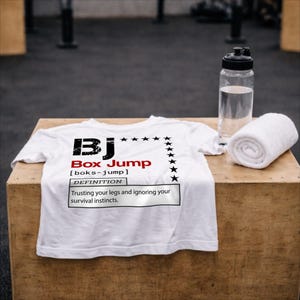 Könnte beinhalten: Weißes T-Shirt mit dem Text "BJ Box Jump" in Schwarz und Rot sowie einer Definition. Eine Wasserflasche und ein gerolltes weißes Handtuch liegen neben dem Shirt. Das Shirt liegt auf einer Holzkiste.