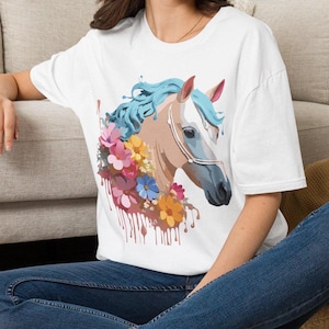 Könnte beinhalten: Ein weißes kurzärmeliges T-Shirt mit einem Grafikdruck eines beigefarbenen Pferdes mit fließender hellblauer Mähne. Das Pferd ist von bunten rosa, gelben und blauen Blumen umgeben, wobei farbähnliche Tropfen vom Blumenmotiv nach unten verlaufen.