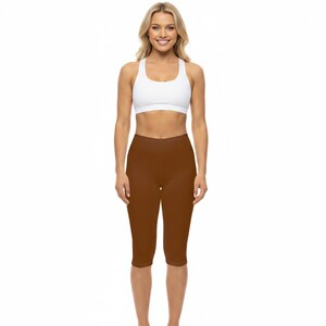 May include: A woman wearing a white sports bra and brown capri leggings. The leggings are a solid, dark brown color. The sports bra is white and has a scoop neck. The woman is smiling and looking at the camera. The background is white.