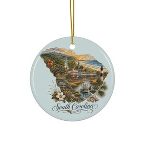 May include: A round, light blue ceramic ornament featuring a detailed illustration of the state of South Carolina. The design includes a coastal landscape, a lighthouse, a ship, peaches, and a white flower. The text South Carolina is printed below.