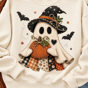 May include: A cream-colored sweatshirt featuring a central graphic of a ghost wearing a black witch hat with stars. The ghost holds an orange pumpkin and is surrounded by black bats, small stars, and a heart. The base of the ghost has patchwork patterns.
