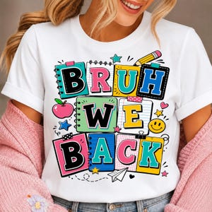 以下が含まれることがあります： 「BRUH WE BACK」という文字がプリントされたカラフルなグラフィックデザインの白い半袖Tシャツ。デザインには、スパイラルノート、鉛筆、ピンクのリンゴ、黄色いスマイリーフェイス、星、紙飛行機のイラストが描かれています。