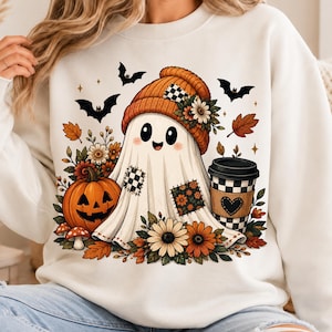Op de afbeelding: Een wit sweatshirt met een afbeelding van een lachende geest die een oranje muts met een bloem draagt. De geest is omringd door herfstbloemen, een pompoen, vleermuizen en een koffiebeker met een schaakbordpatroon en een zwart hartje.