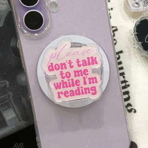 Puede incluir: Una funda de teléfono con purpurina lavanda con un soporte para teléfono que dice "Please don't talk to me while I'm reading". También se ve un lector Kindle y pines en forma de corazón con texto.
