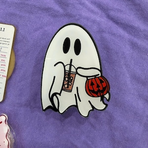 May include: A white ghost-shaped pin with black eyes and a black outline. The ghost is holding a pumpkin and a drink with a straw. The pin is on a purple background. Halloween-themed.
