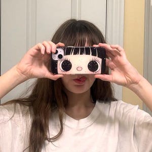 Op de afbeelding: Zwart-witte telefoonhoes met een cartoonmeisje met grote ogen en wimpers. De hoes heeft een zwarte achtergrond met een wit gezicht en grote zwarte ogen. De telefoonhoes wordt voor het gezicht van een persoon gehouden.