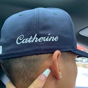 May include: A navy blue baseball cap featuring the name Catherine embroidered in white script lettering on the side panel. The cap has a structured crown, a stiff brim, and a small white rectangular tag on the lower edge near the back.