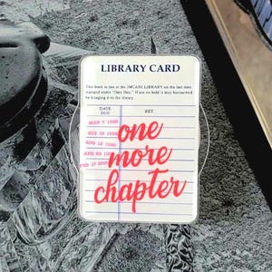May include: A Kindle e-reader with a clear case displays a grayscale image. A sticker in the shape of a library card is in front of the Kindle, with the words "one more chapter" in red. Halloween-themed pins surround the e-reader.