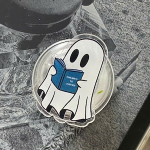 Puede incluir: Una pegatina blanca con forma de fantasma leyendo un libro azul con el texto "Enemies to Lovers" se coloca en un lector de libros electrónicos Kindle. El lector es negro con una pantalla blanca que muestra una imagen en escala de grises. El fondo es una superficie gris claro.
