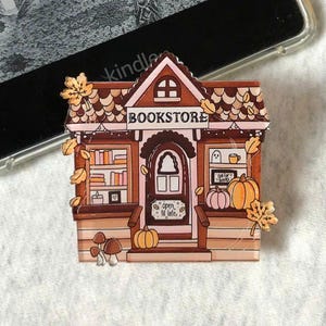 May include: A bookstore-themed pin in brown and tan tones. The pin depicts a detailed illustration of a bookstore with pumpkins, leaves, and the word "BOOKSTORE" on the roof. The pin also has the words "Open 'til late" on the door.