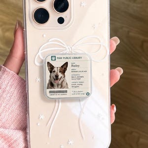 Puede incluir: Funda transparente para teléfono con diseño de perro. La funda presenta una foto de un perro llamado Bailey, un lazo blanco y detalles de estrellas. El texto "Paw Public Library" es visible.