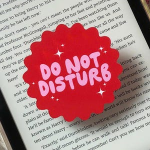 May include: A red, scalloped-edge sticker with the words "DO NOT DISTURB" in pink, with white starbursts. The sticker is on a Kindle screen displaying book text.