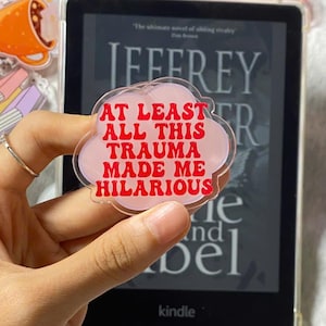 Puede incluir: Un charm de acrílico en forma de nube rosa con el texto "AT LEAST ALL THIS TRAUMA MADE ME HILARIOUS" en rojo. El charm se sostiene frente a un lector Kindle. Otros charms decorativos son visibles en el fondo.