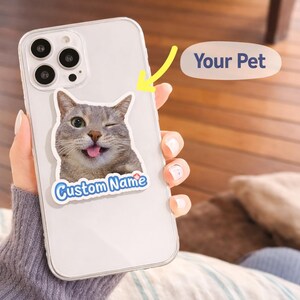 Puede incluir: Una funda transparente para teléfono con una pegatina que muestra un gato guiñando un ojo y sacando la lengua. La pegatina tiene el texto "Custom Name" en azul y blanco. La imagen también incluye una burbuja de texto que dice "Your Pet".