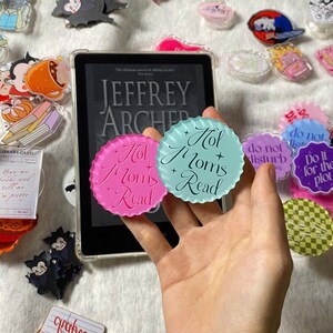 May include: A hand holds two round, scalloped-edge acrylic book markers. One is pink, the other is light blue, both with the text "Hol Moms Read" in script. Other book-themed accessories and a book are in the background.