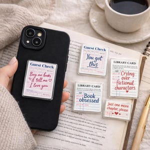 May include: A black phone case with a clear phone grip featuring a "Guest Check" design with the text "Buy me books & tell me I love you." Several other clear stickers with book-themed text are scattered around.