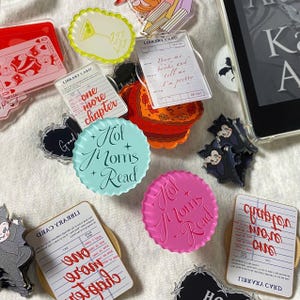 May include: A collection of colorful acrylic charms and accessories, including library card designs, bat-shaped figures, and round designs with the text "Hot Moms Read." Other charms feature text like "One More Chapter" and "Buy me books and tell me I'm pretty."