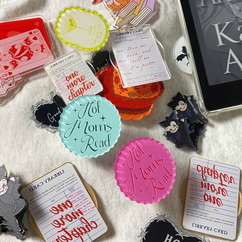 May include: A collection of colorful acrylic charms and accessories, including library card designs, bat-shaped figures, and round designs with the text "Hot Moms Read." Other charms feature text like "One More Chapter" and "Buy me books and tell me I'm pretty."