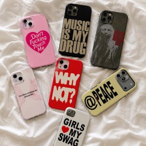 May include: A collection of seven smartphone cases featuring various designs: a pink case with a heart and text, a black case with gold text, a dark grey case with a portrait, a white case with pink watercolour, a red case with white text, a yellow case with black text, and a white case with black text.