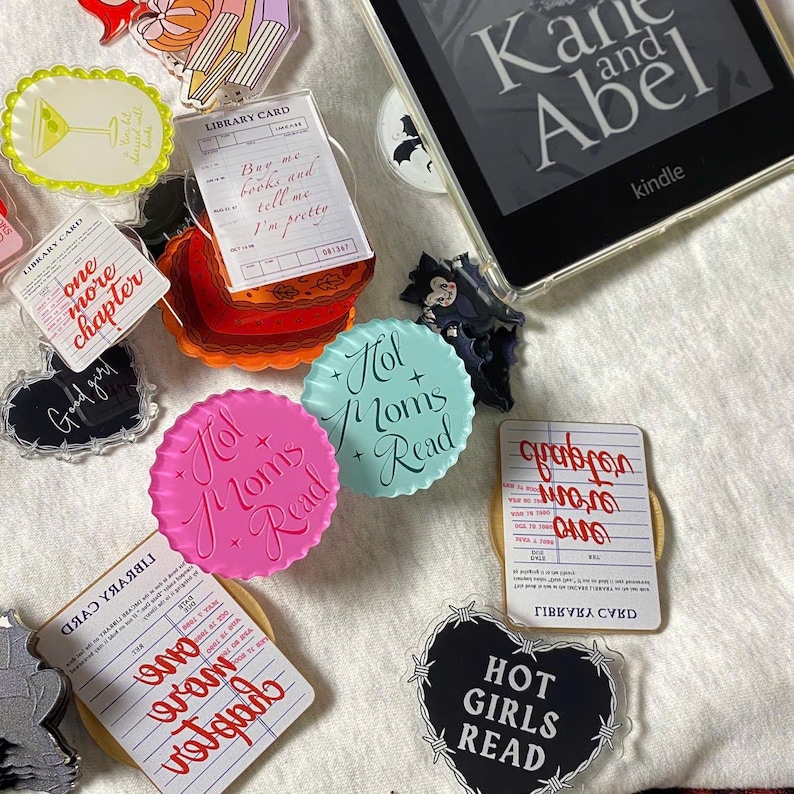May include: A collection of colorful acrylic book accessories, including library card and heart-shaped designs with text like "Hot Moms Read" and "Hot Girls Read." A Kindle e-reader displays the title "Kane and Abel."