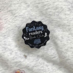 May include: A black and white Fantasy Readers Book Club pin. The pin has a scalloped edge and features the text "Fantasy readers Book Club" in white and light blue. The pin also has illustrations of stars, bats, and books.