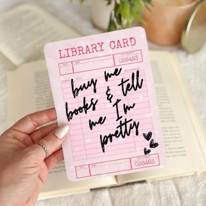 May include: A pink library card with the words "buy me books & tell me I'm pretty" in black script. The card has a vintage design with lines and the words "LIBRARY CARD" at the top. Three small black hearts are on the right side.