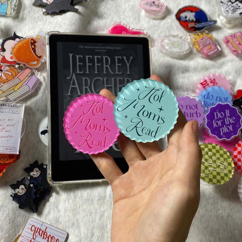 May include: A hand holds two round, scalloped-edge acrylic pins. One is pink with "Hot Moms Read" in script, the other is light blue with the same text. Other pins and a tablet with a book cover are in the background.