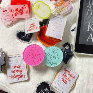 May include: A collection of acrylic charms and accessories with bookish and Halloween themes. Includes pink and teal circular charms that read "Hot Moms Read", library card designs, and a playing card design. Other charms have text like "Buy me books & tell I'm pretty."