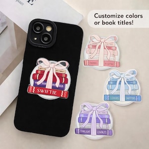 Op de afbeelding: Een zwarte telefoonhoes met een decoratieve sticker met een stapel boeken vastgebonden met een roze strik. De sticker zegt "Swiftie" en "Why Liv Butthees". Extra stickers tonen stapels boeken met de tekst: "Enemies to Lutters", "Lovers Shitfiers" en "Starlight Loverless".