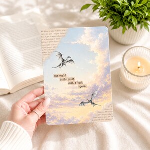 Puede incluir: Un cuaderno rectangular con un diseño de dragón y nubes. El texto en el cuaderno dice: "The world falls quiet when a book opens." El cuaderno está sostenido en una mano, con un libro abierto y una vela encendida en el fondo.