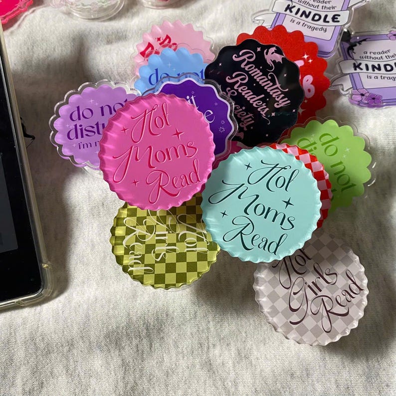 May include: A collection of colorful, decorative pop sockets with various designs and text. The pop sockets feature phrases like "Hot Moms Read" and "Romantasy Readers Society". Colors include pink, blue, green, and black.