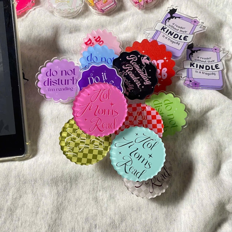 May include: A collection of colorful, circular acrylic book accessories with various phrases, including "Hot Moms Read," "Do Not Disturb," and "A reader without their Kindle is a tragedy."