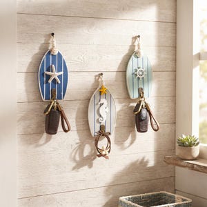 May include: Three surfboard-shaped wall hooks featuring vertical blue and white stripes. The left hook has a white starfish, the centre has a yellow starfish and white seahorse, and the right has a white ship wheel. Each includes a rope hanger.