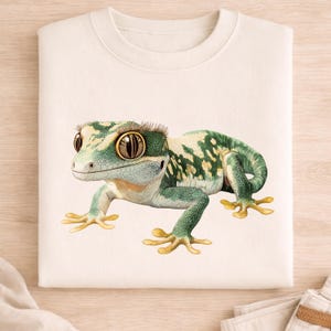 Può includere: Felpa color crema con un'illustrazione dettagliata di un geco. Il geco ha un motivo a macchie verdi e bianche, grandi occhi marroni e piedi gialli. Il disegno è centrato sulla parte anteriore della felpa.