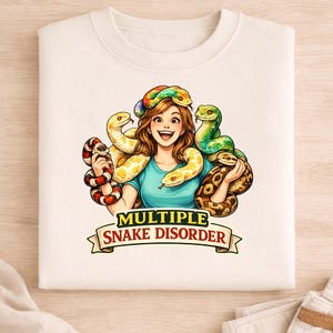 May include: A cream-coloured sweatshirt with a cartoon illustration of a smiling person surrounded by colourful snakes. The text "MULTIPLE SNAKE DISORDER" is displayed on a banner below the illustration.