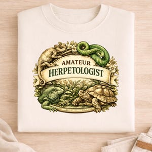 Può includere: Felpa color crema con un'illustrazione dettagliata di un camaleonte, un serpente, un'iguana e una tartaruga. Il design include il testo "AMATEUR HERPETOLOGIST" su uno striscione, circondato da fogliame. Una maglietta grafica unica per gli appassionati di rettili.