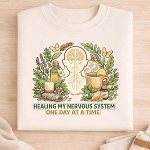 Può includere: Felpa color crema con una grafica raffigurante una testa umana stilizzata con un design del cervello, circondata da piante, farfalle e una tazza di tè. Il testo recita: "HEALING MY NERVOUS SYSTEM ONE DAY AT A TIME."