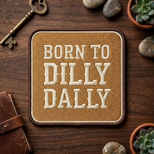 May include: Square patch with the embroidered text "BORN TO DILLY DALLY" in white on a tan background. The patch has a brown border and is surrounded by a key, small succulents, and a leather-bound notebook on a wooden surface.