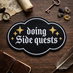 May include: Black embroidered patch with the words "doing Side quests" in white gothic lettering, surrounded by gold stars. The patch is on a wooden surface with a compass, key, and other items.